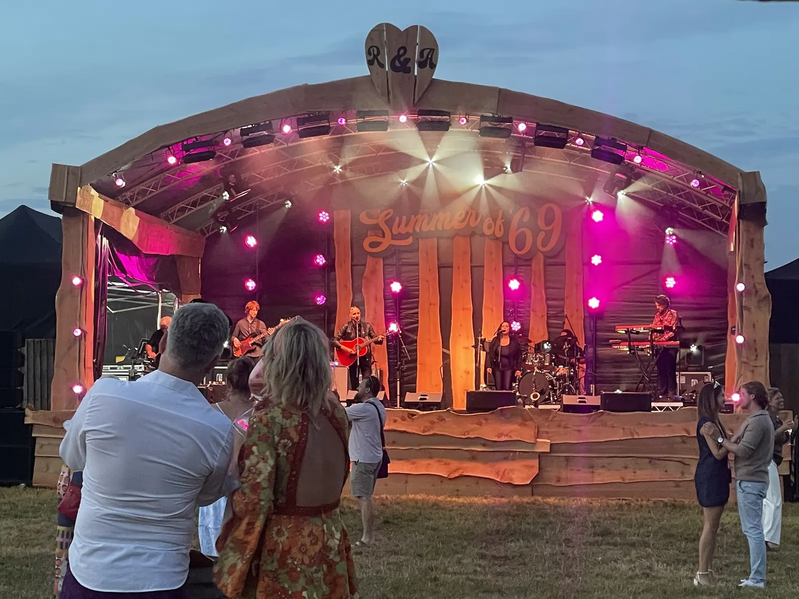 An outdoor festival-style wooden stage at a private wedding party with a live band, roof truss and pink lighting at dusk